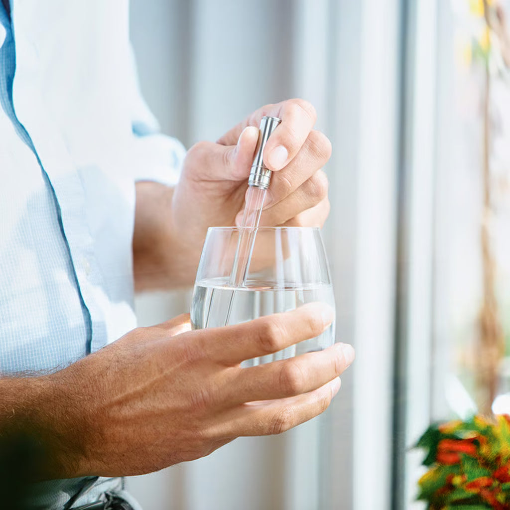 Hånd som holder et glass med klart vann, mens en sølvfarget pipette senkes ned i vannet.