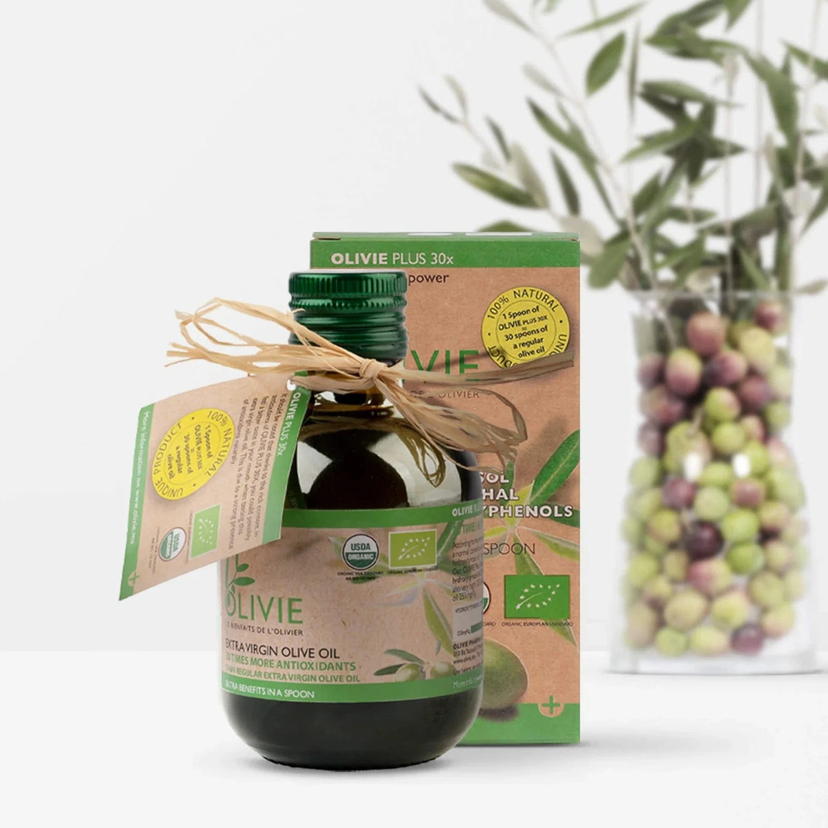 Olivenolje i mørk glassflaske med grønn plastlokk, merket OLIVIE Plus 30x, med bånd og etikett. Bakgrunn med oliven.