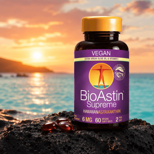 BioAstin Supreme astaxantin i en brun plastflaske med gul lokk, seks røde softgels på svart stein, horisont med solnedgang.