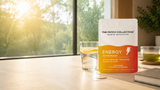 Wellnessplasteret "Energy Patch" står på et bord ved vinduet. Plasteret har en fargerik emballasje med gul og oransje design, og det er en liten vannfylt drikkeglass med sitron på bordet.