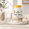 GABA 750 mg kapsler i hvit emballasje med gultt lokk, plassert ved et glass med vann og en steinplate.