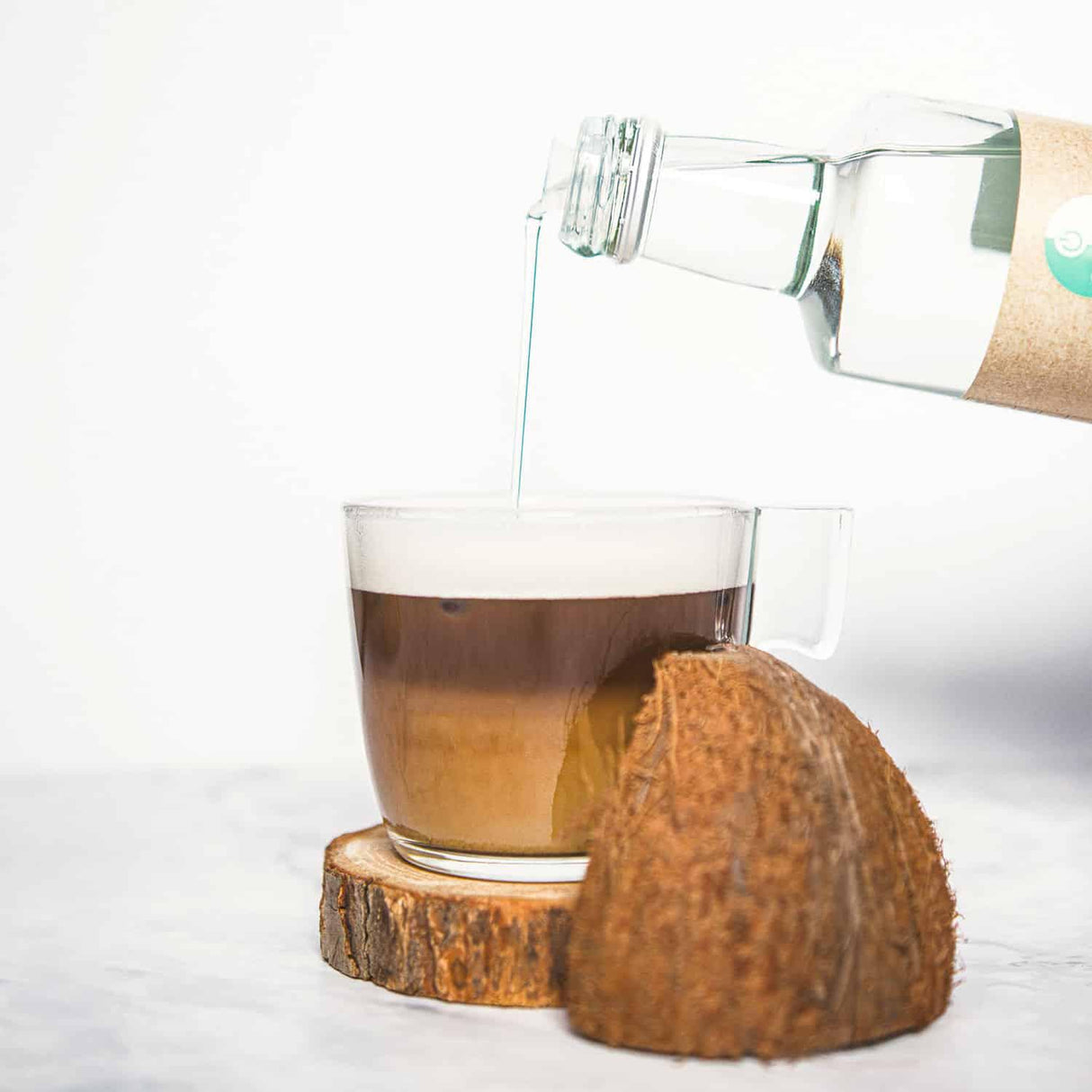 GoKeto Bio MCT-olje helles fra en glassflaske i en kopp med kaffefarge og skum på toppen, med en kokosnøtt ved siden av.