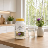 Mariendistel Max-flaske med gult lokk står på et trebord ved siden av et glass med vann og en vase med blomster.